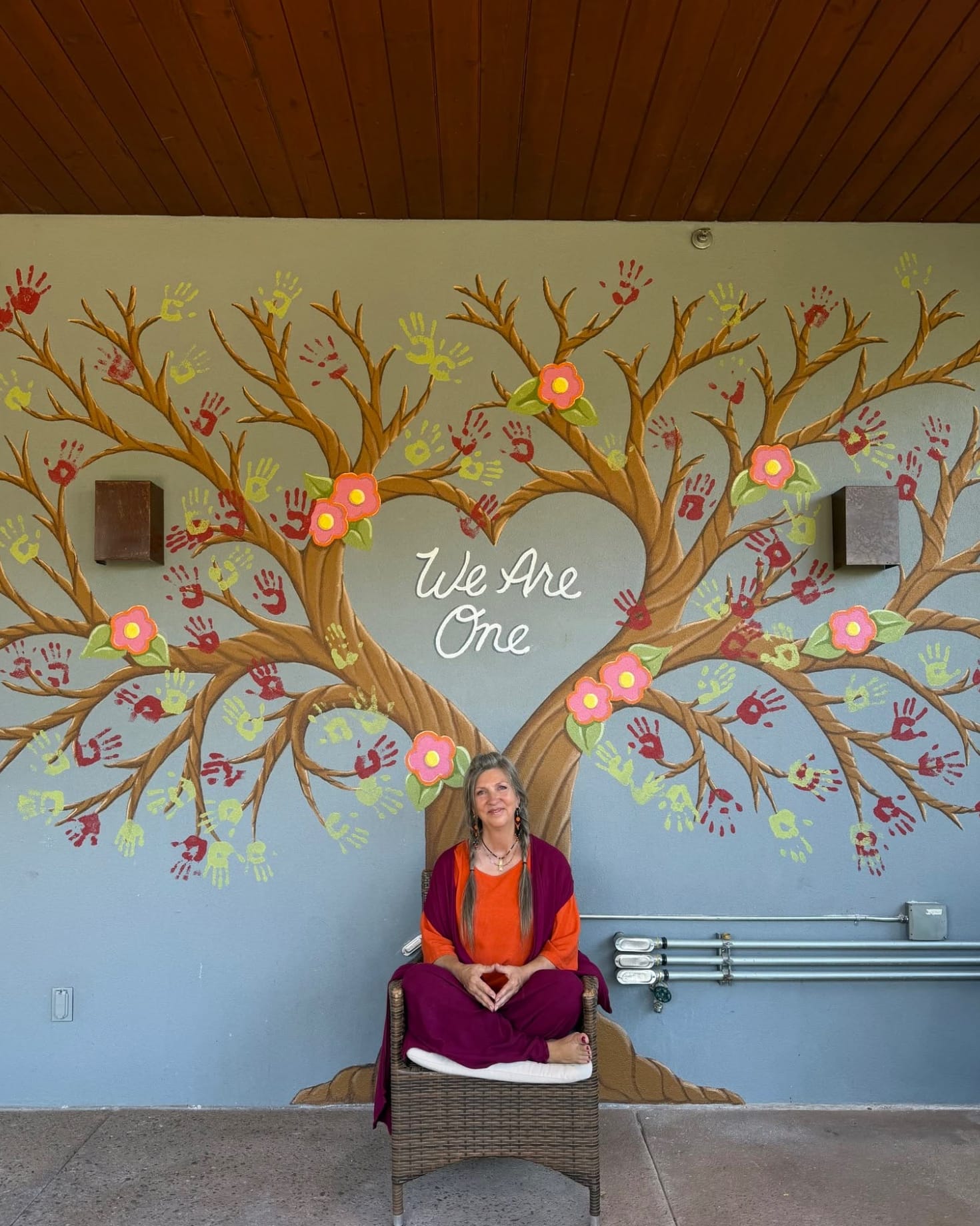 A teacher seated before a heart-shaped tree mural reading 'We Are One,' its branches covered in community handprints