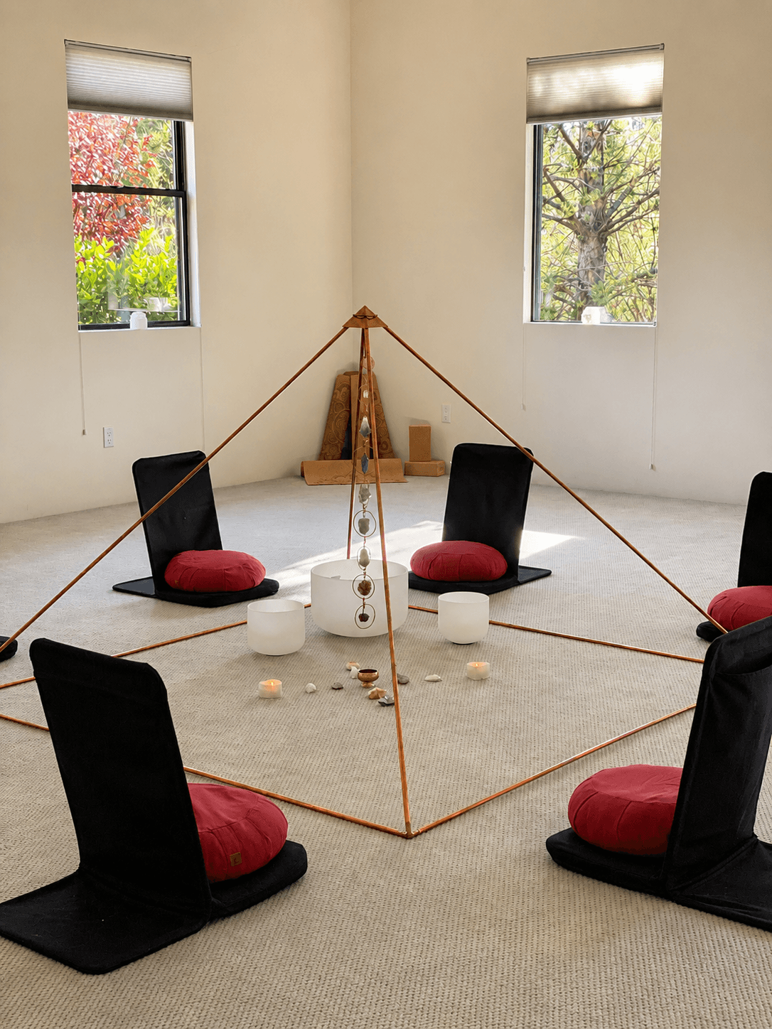 A copper pyramid frame surrounded by cushioned meditation chairs, singing bowls and candles set at its center, beneath Sicatuva's tall windows